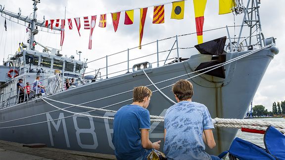 Twee jongens zitten aan de kade bij een schip van de Marine