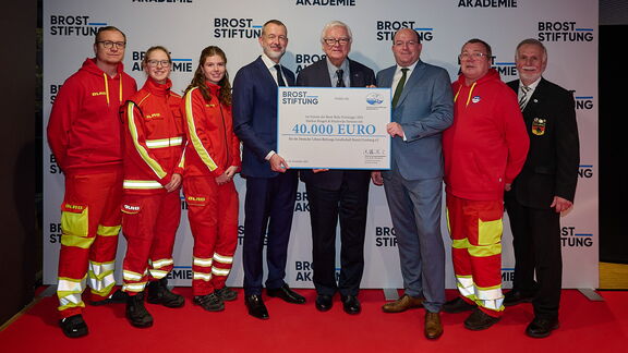 Eine Gruppe von Personen steht auf einem roten Teppich vor einer Wand mit dem Logo „Brost Stiftung“ und „Akademie“. Mehrere Personen tragen rote Rettungsuniformen mit gelben Reflexstreifen, andere sind in formeller Kleidung. In der Mitte wird ein großer symbolischer Scheck über 40.000 Euro präsentiert.