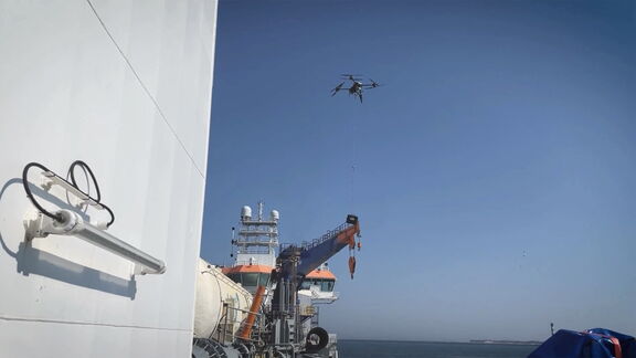Eine Drohne schwebt über einem Schiff auf offener See und lässt ein Kabel zu einem Deckkran herab, während Radarkuppeln und Antennen vor einem klaren blauen Himmel sichtbar sind.