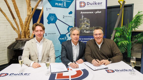 Three people sit at a table signing documents during a formal event at Duiker Clean Technologies, with company banners and glasses of champagne in the background.
