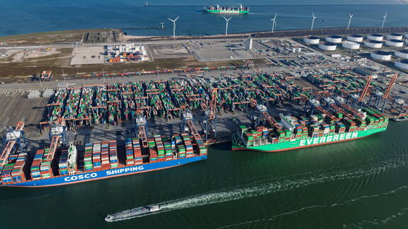 Luftaufnahme des RWG‑Terminals mit mehreren großen Containerschiffen, darunter COSCO SHIPPING und EVERGREEN, die am Kai liegen. Zahlreiche Kräne verladen Container, während im Hintergrund ein weitläufiges Gelände mit gestapelten Containern und Windturbinen an der Küste zu sehen ist.