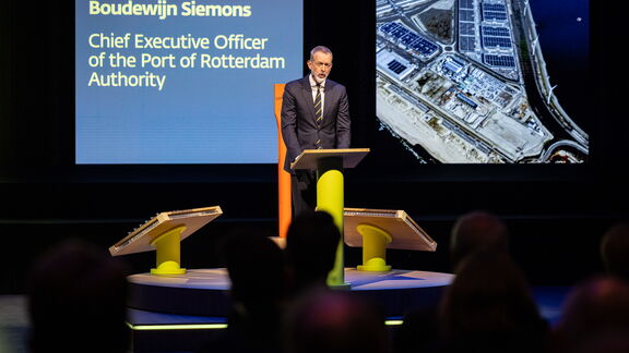 Spreker staat achter een podium op een conferentie, met een presentatie over de haven van Rotterdam op de achtergrond.