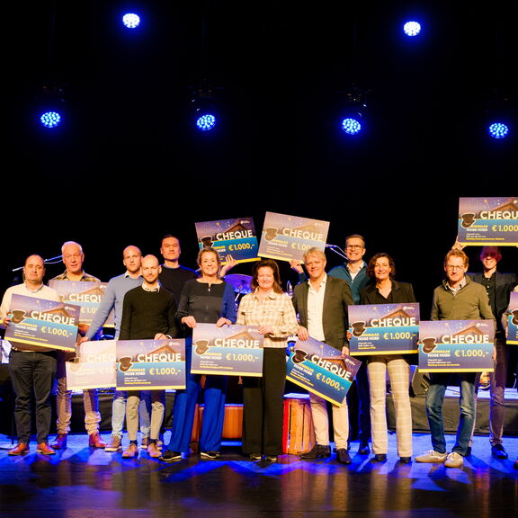 Groep mensen op een podium tijdens een prijsuitreiking, elk houdt een grote cheque van €1.000 vast. Het podium is verlicht met blauwe spotlights en de achtergrond is donker, waardoor de cheques duidelijk zichtbaar zijn.