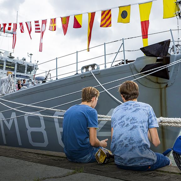 Twee jongens zitten aan de kade bij een schip van de Marine
