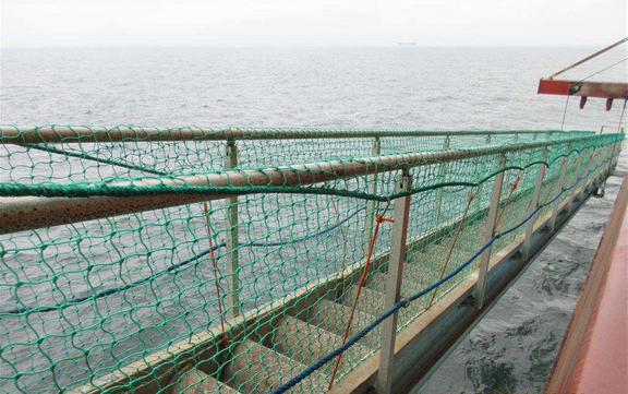 Een scheepsdek met netten langs de reling boven een ruwe zee, gezien vanaf de zijkant van het schip