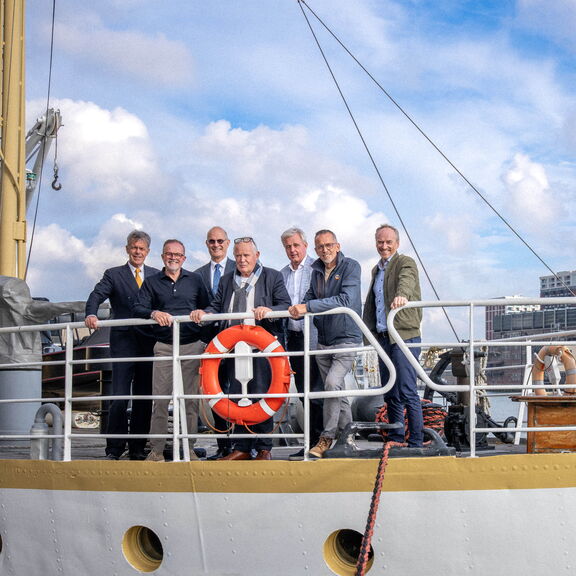 Een groep vertegenwoordigers staat op het dek van een schip,poserend voor een foto met een reddingsboei op de voorgrond en havengebouwen op de achterground.