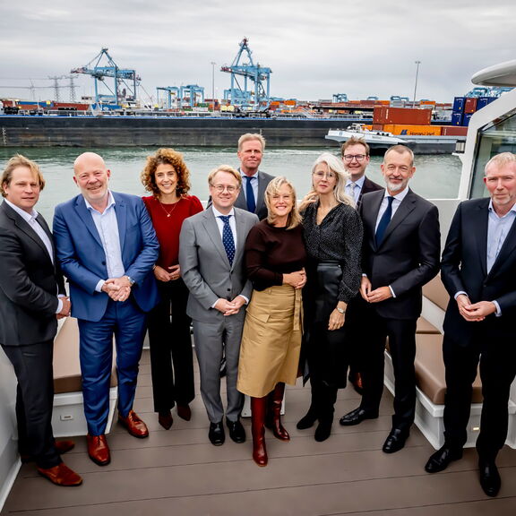 Een groep mensen staat samen op het dek van een boot in een havenomgeving. Op de achtergrond zijn containers, kranen en schepen zichtbaar, wat wijst op een industriële havenlocatie. De personen zijn netjes gekleed in zakelijke kleding, waaronder pakken en formele outfits. Het water en enkele boten zijn duidelijk te zien, evenals een moderne inrichting van het dek.