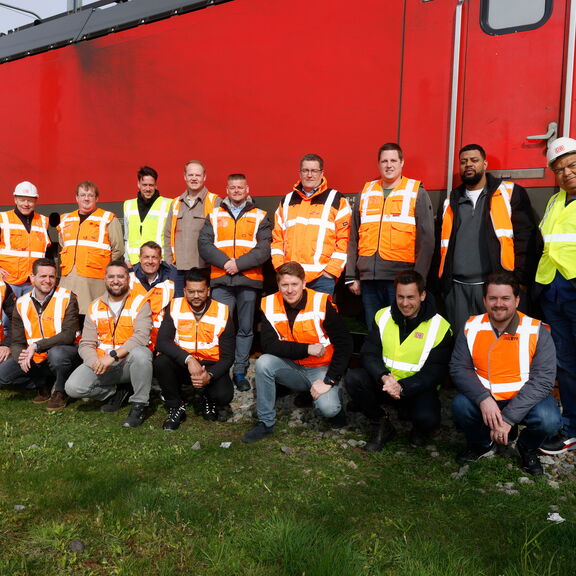 „Gruppenfoto von etwa zwanzig Personen in orangefarbenen und gelben Sicherheitswesten, die vor einem roten Eisenbahnwaggon posieren. Sie stehen auf Gras und Schotter, einige hockend, andere stehend, alle in die Kamera blickend.